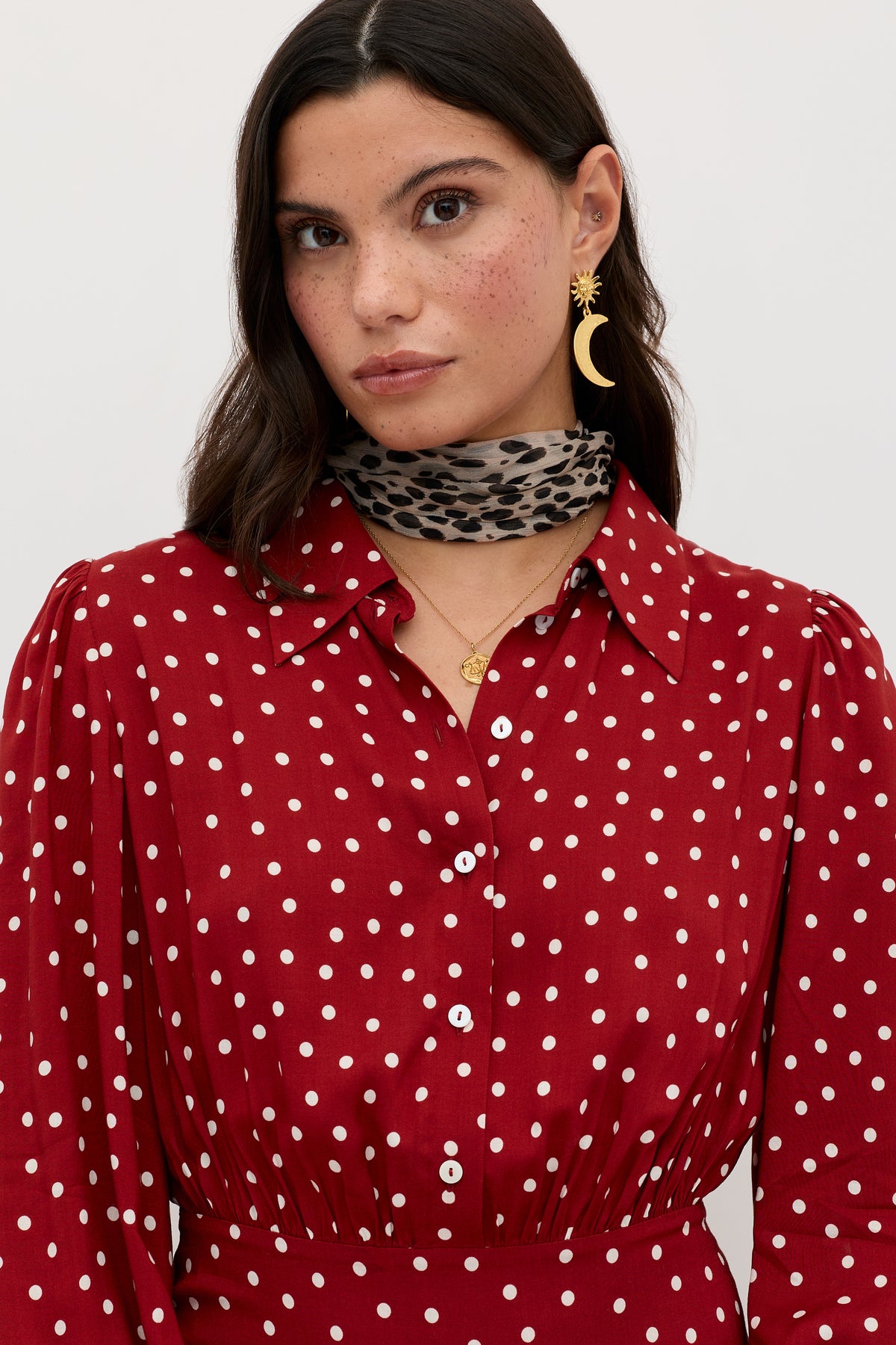 A woman with long dark hair wears the Izzy - Layla Dot Red dress, featuring white polka dots, paired with a leopard print scarf, gold crescent moon earrings, and a gold necklace against a plain light background.