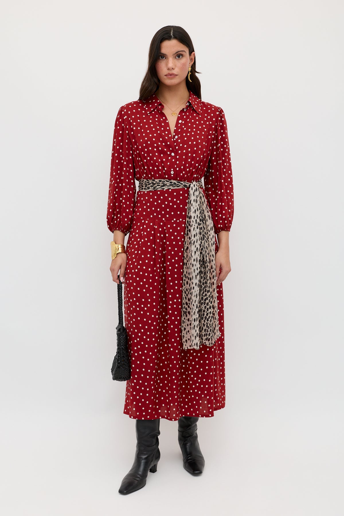 A woman stands against a white background wearing the Izzy - Layla Dot Red dress, paired with black boots, a black textured bag, a gold bracelet, and a patterned scarf tied at her waist.