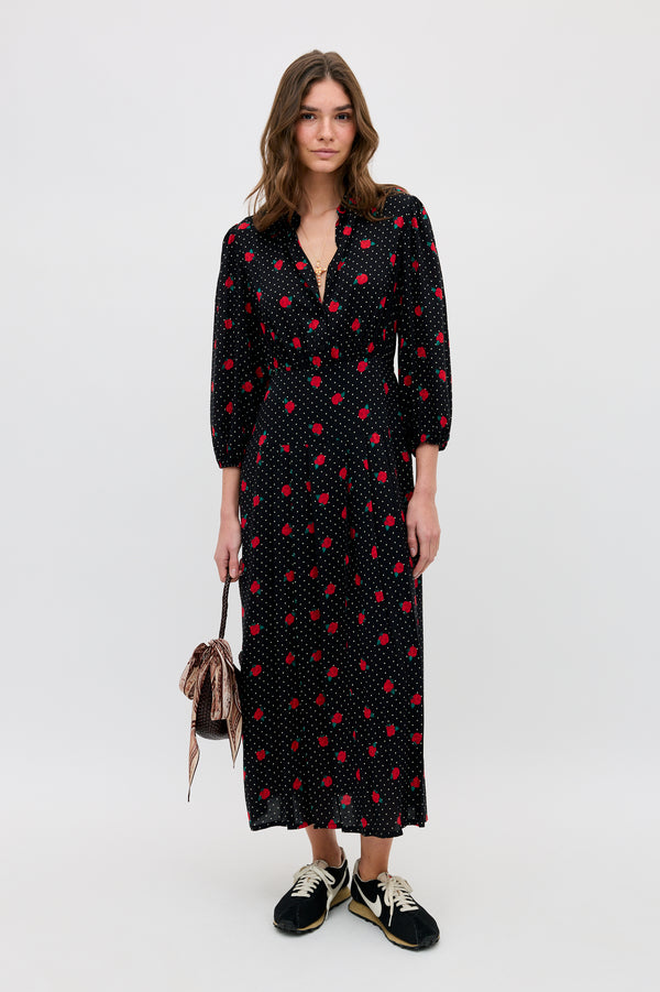 A woman with wavy hair and a relaxed expression stands against a plain background wearing the Izzy - Rose Spot Black long dress with red polka dots, black sneakers, and holding a small striped drawstring bag.