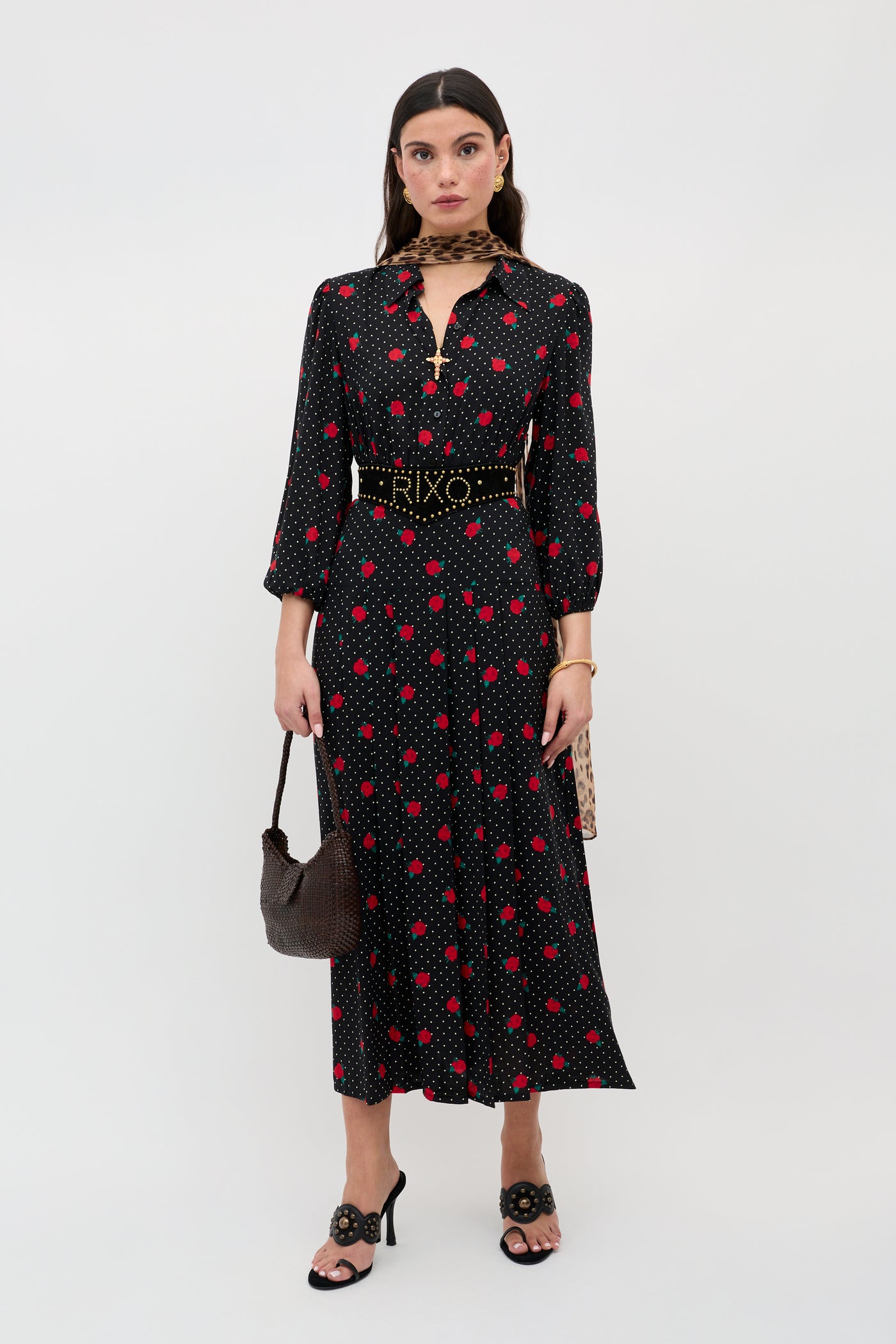 A woman stands against a plain background wearing the Izzy - Rose Spot Black dress, an ankle-length, black long-sleeve dress with red and white polka dots, paired with a gold-buckled belt, black sandals, a brown handbag, and a patterned scarf.