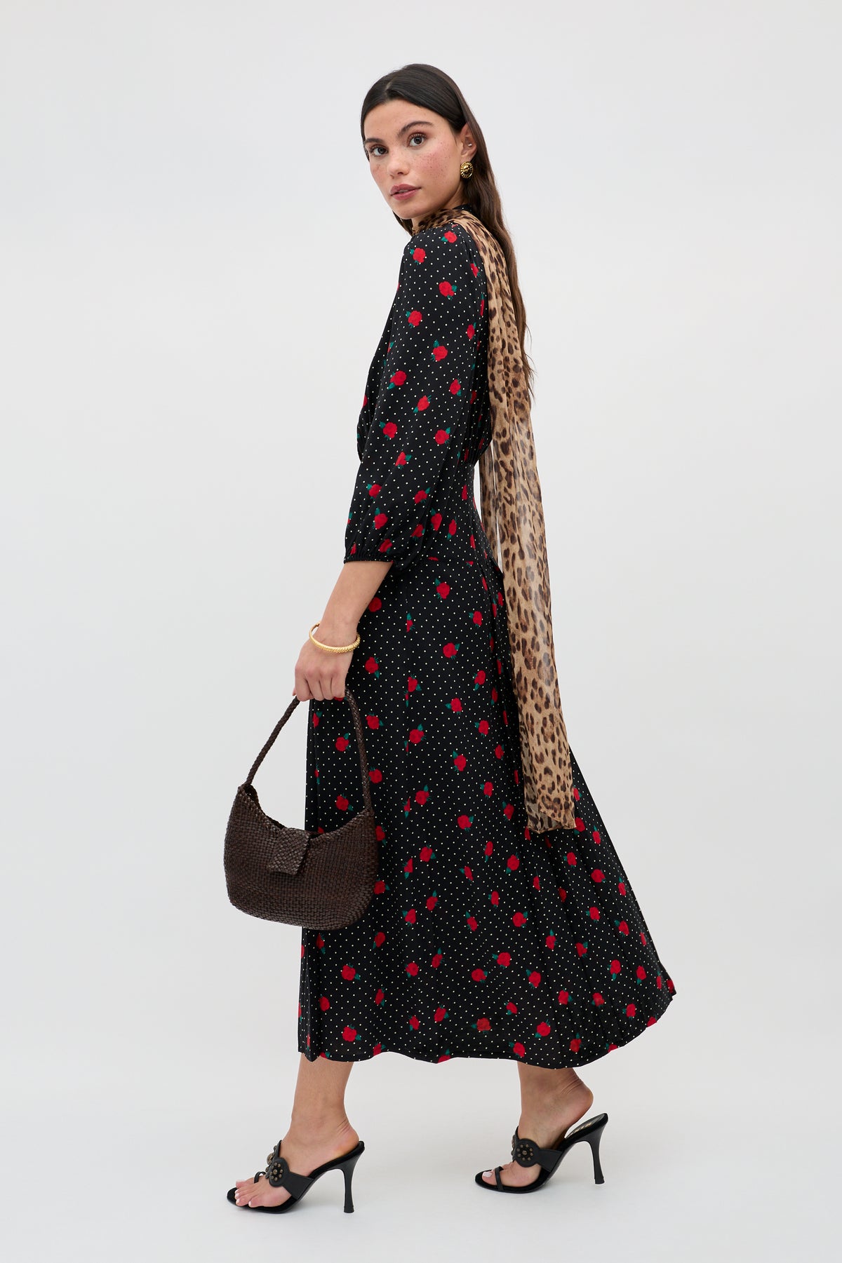 A woman wears the Izzy - Rose Spot Black long-sleeved, ankle-length dress with red polka dots, paired with black heeled sandals, a brown woven handbag, and a leopard print scarf, posing against a plain light background.