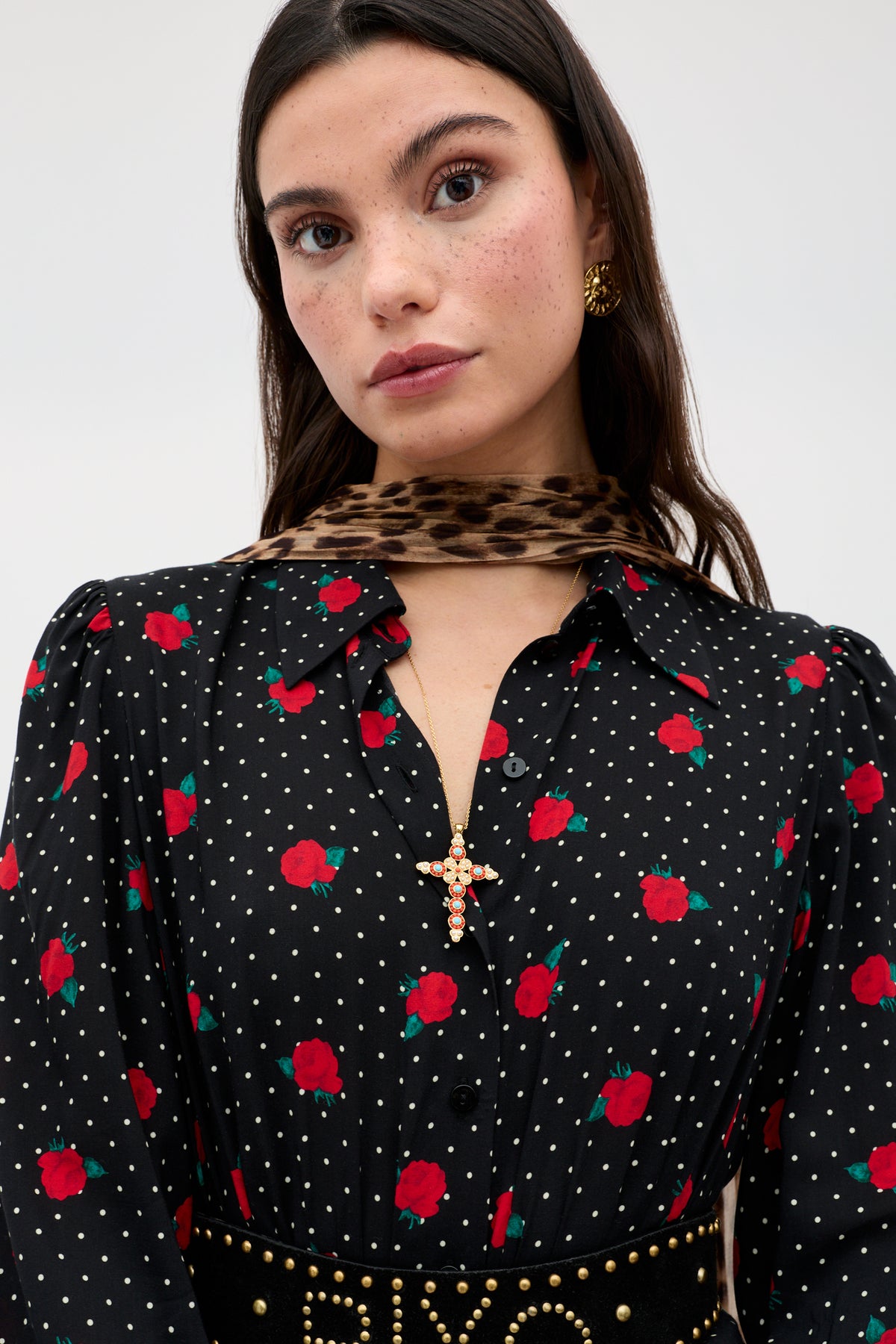 A woman with long brown hair wears the Izzy - Rose Spot Black dress, featuring red rose and white polka dot patterns, styled with a leopard print scarf, gold earrings, and a cross-shaped pendant necklace.