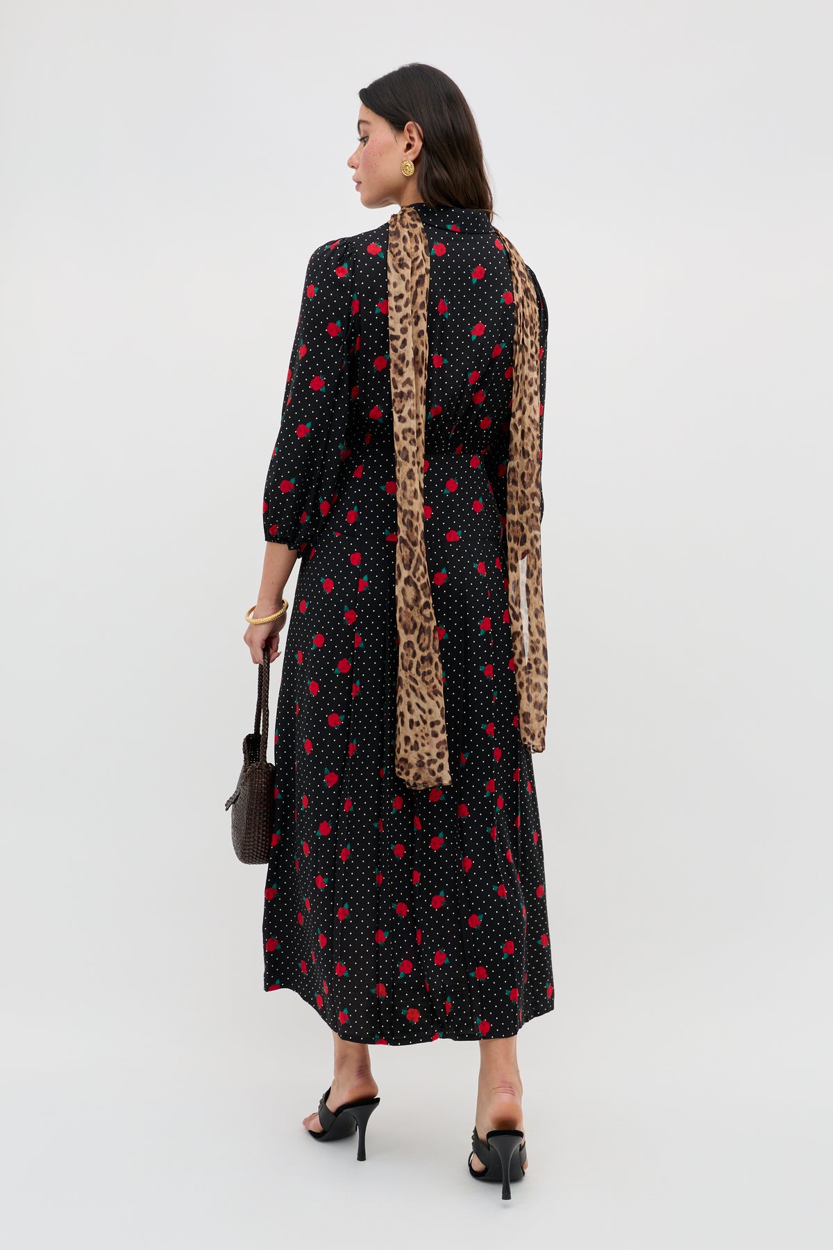 A woman wearing the Izzy - Rose Spot Black long-sleeved, ankle-length dress stands facing away, styled with black heels, a long leopard-print scarf, and a brown handbag.