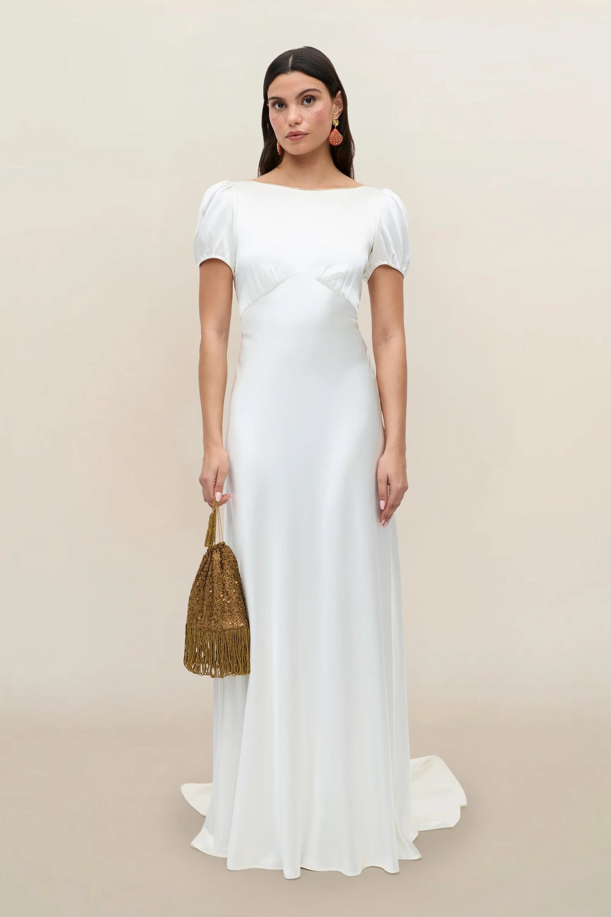 A woman stands against a plain background wearing the Natalia gown in satin back silk crepe, a white thirties-inspired dress with short sleeves. She holds a gold tasseled handbag and accessorizes with earrings featuring a subtle diamanté trim. Her long, dark hair is worn loose, and she gazes directly at the camera with a neutral expression.
