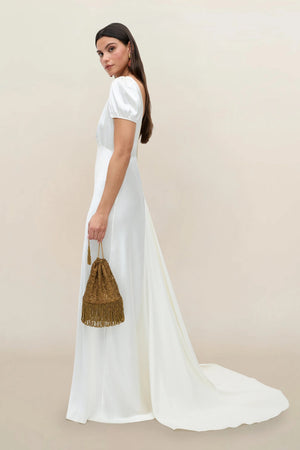 A woman in the Natalia satin back silk crepe gown, inspired by 1930s fashion with short sleeves and a sweeping floor-length train, stands sideways holding a gold tasseled handbag against a plain neutral background.