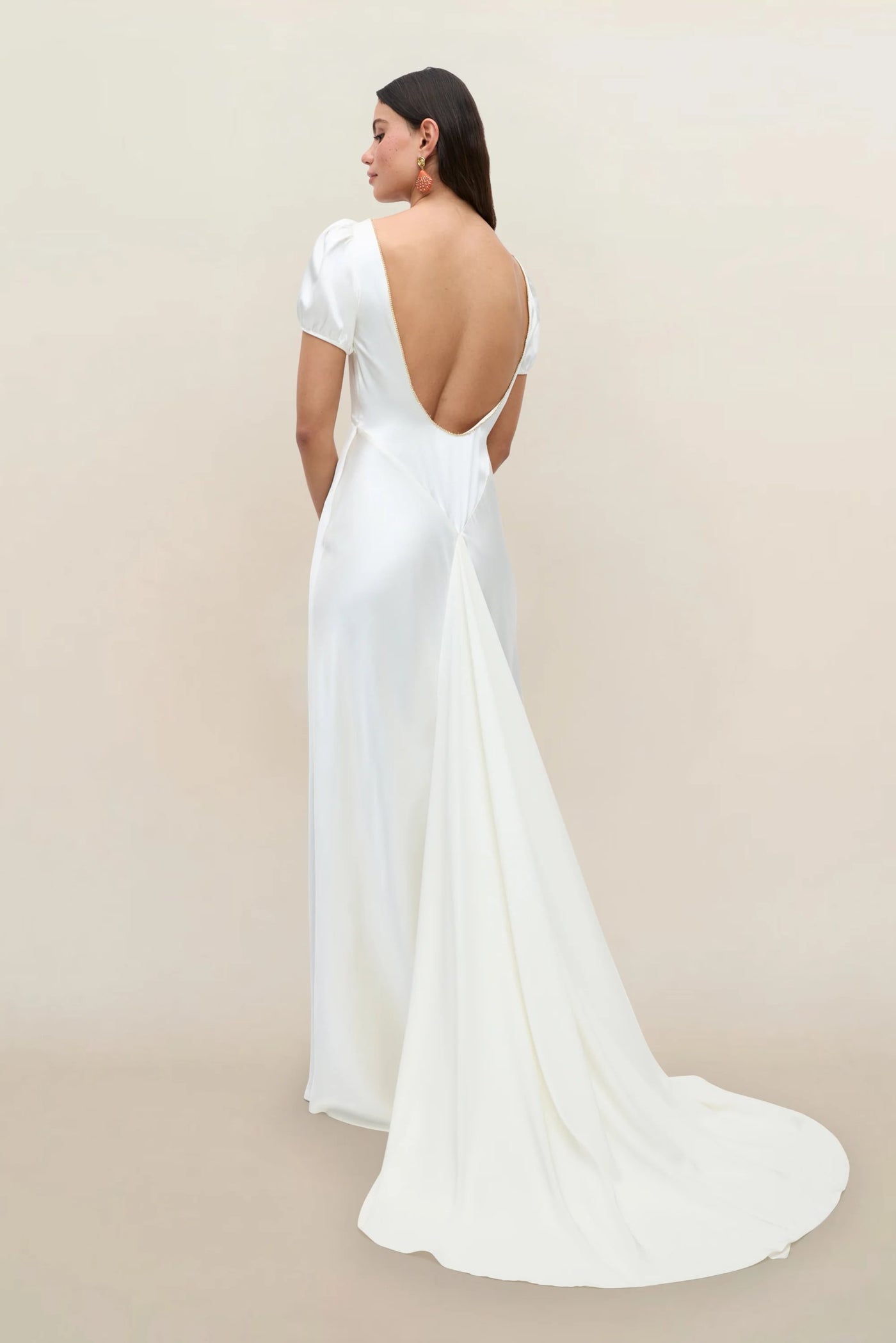 A woman stands against a neutral background, facing away from the camera, wearing the Natalia - Satin Back Silk Crepe gown—a Thirties-inspired, white silk dress featuring a diamanté trim, backless design, short sleeves, and a long, flowing train.