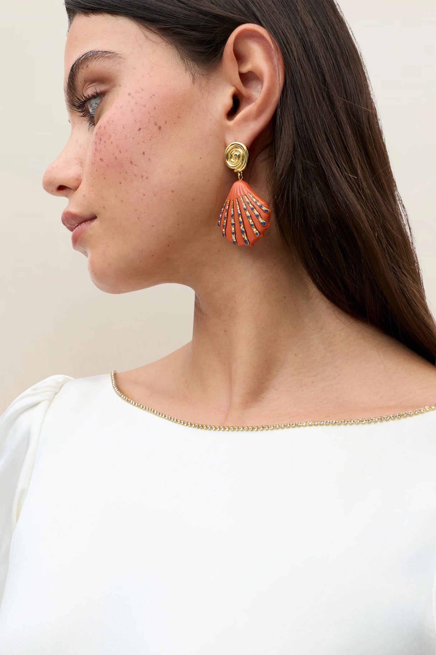 A woman, evoking thirties-inspired elegance in a white Natalia Satin Back Silk Crepe top with gold trim, looks to the left, her look completed by an orange and gold shell-shaped earring with a gold rose stud.