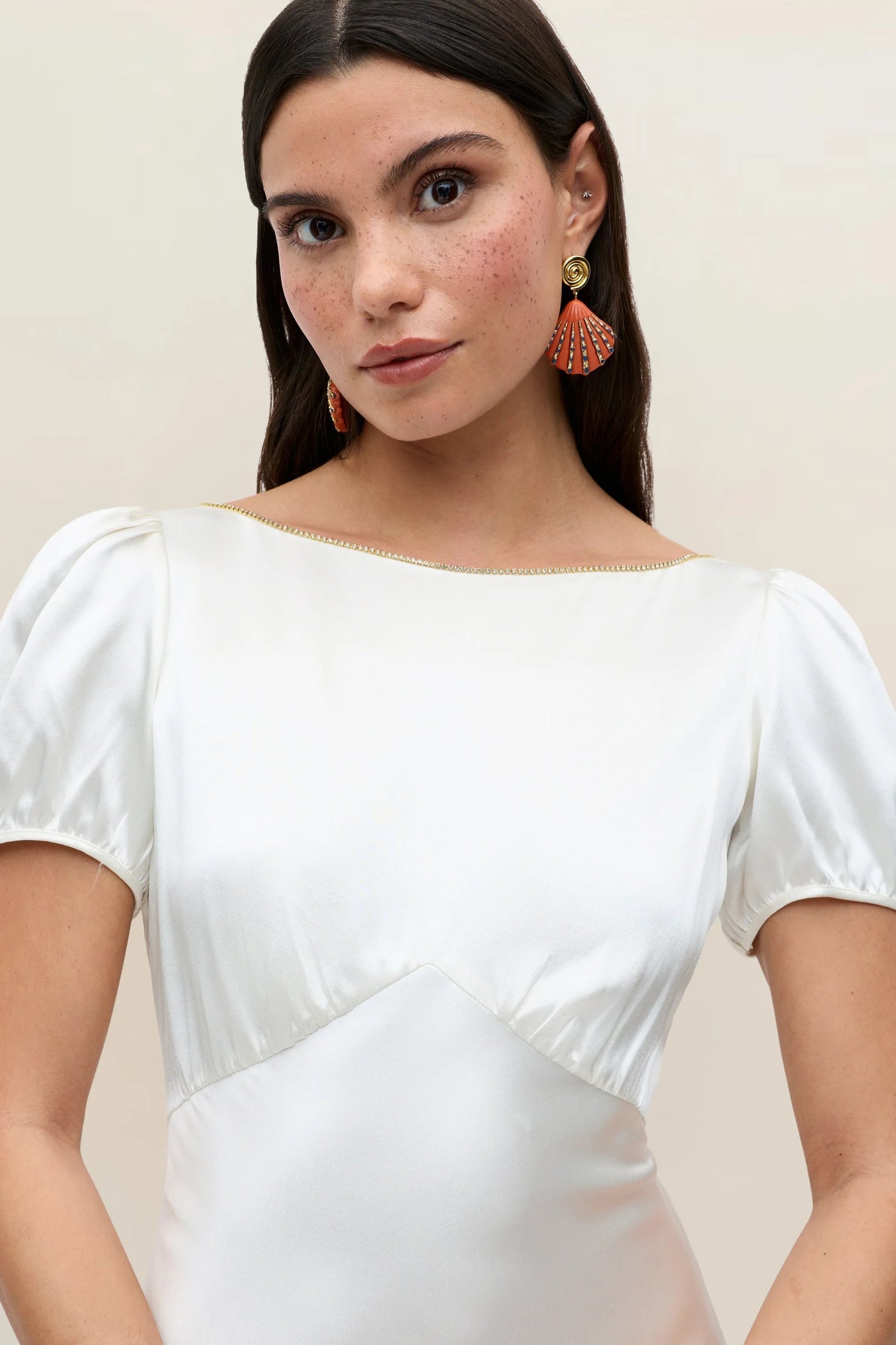 A woman with long brown hair, wearing the Natalia - Satin Back Silk Crepe gown—a Thirties-inspired white silk dress with short puff sleeves—accented by orange fan-shaped earrings and a subtle diamanté trim, looks at the camera against a plain light background.