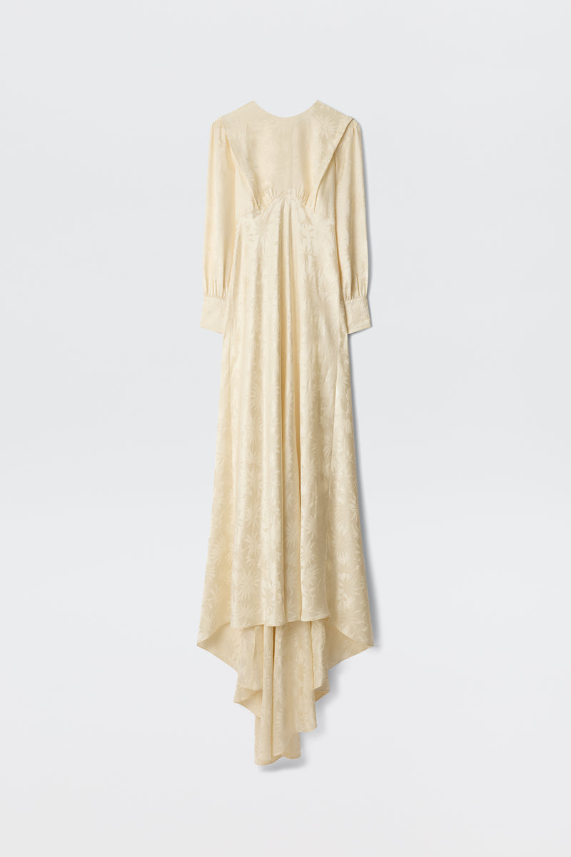 A long champagne-colored Daisy Jacquard gown named “Clementine,” featuring long sleeves, subtle floral patterns, a fitted waist, and an asymmetrical hem that extends longer in the back, displayed against a plain white background.