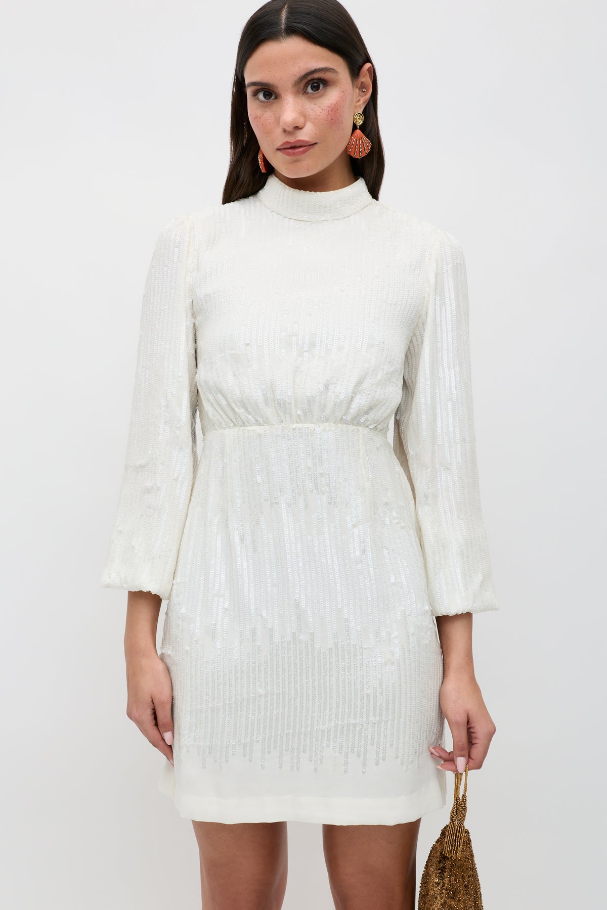 A woman wears the "lara - ivory black bow," a white long-sleeve sequinned mini dress featuring a high neckline and elegant black bow detail. She accessorizes with statement earrings and a small gold bag, standing against a plain white background.
