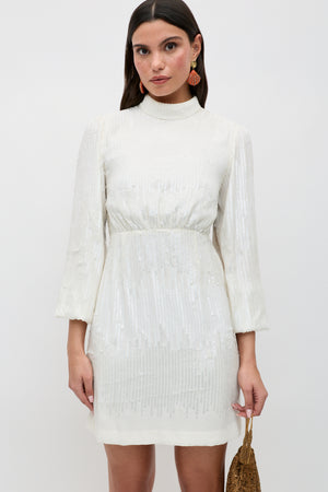 A woman wears the "lara - ivory black bow," a white long-sleeve sequinned mini dress featuring a high neckline and elegant black bow detail. She accessorizes with statement earrings and a small gold bag, standing against a plain white background.