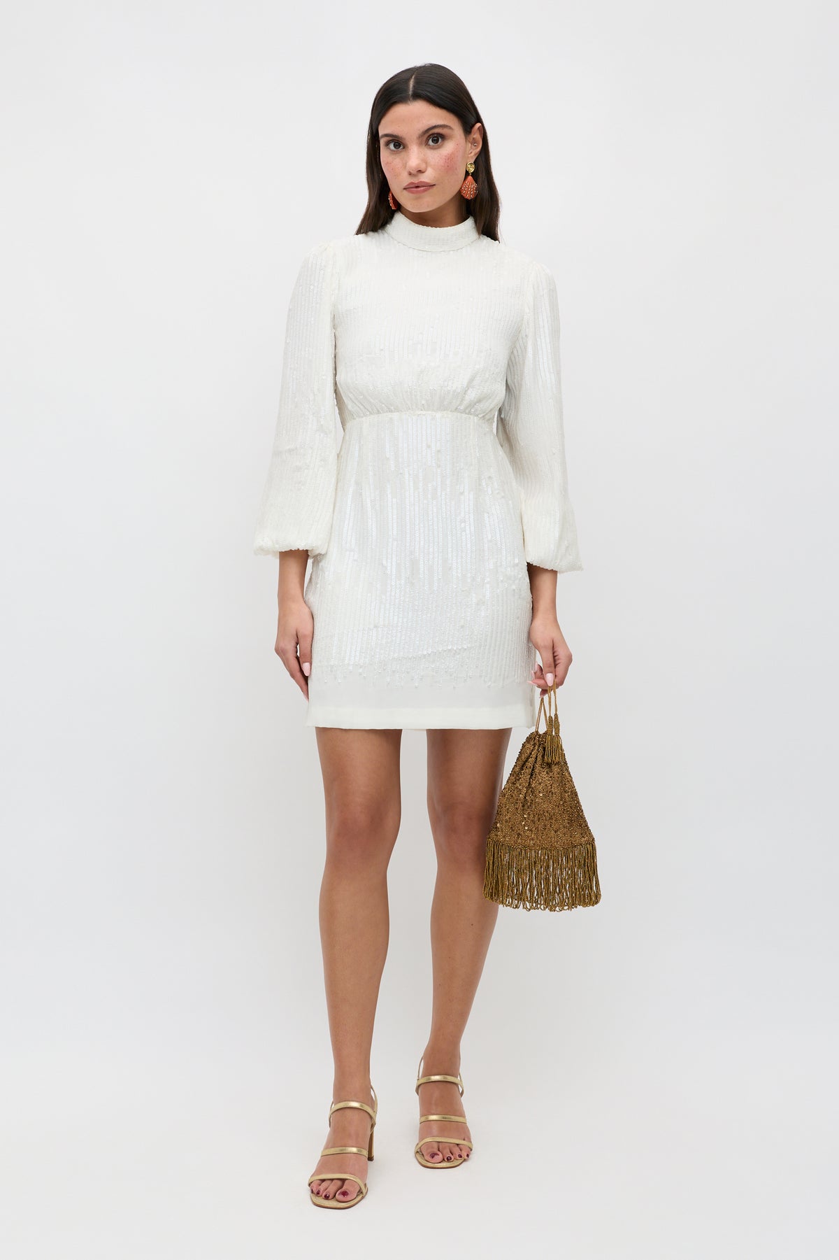 A woman stands against a plain white background wearing the ‘lara - ivory black bow’ dress, a white, long-sleeved, knee-length design featuring a tie-back detail. She accessorizes with large earrings, gold sandals, and holds a gold fringed handbag.