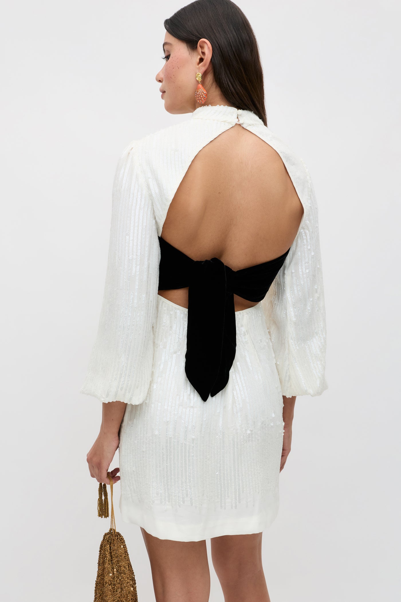 Woman in the "lara - ivory black bow": an ivory, long-sleeved sequinned dress with an open back and black bow detail. She pairs gold earrings with a gold beaded handbag—a stunning tie-backed ensemble perfect for elegant occasions.