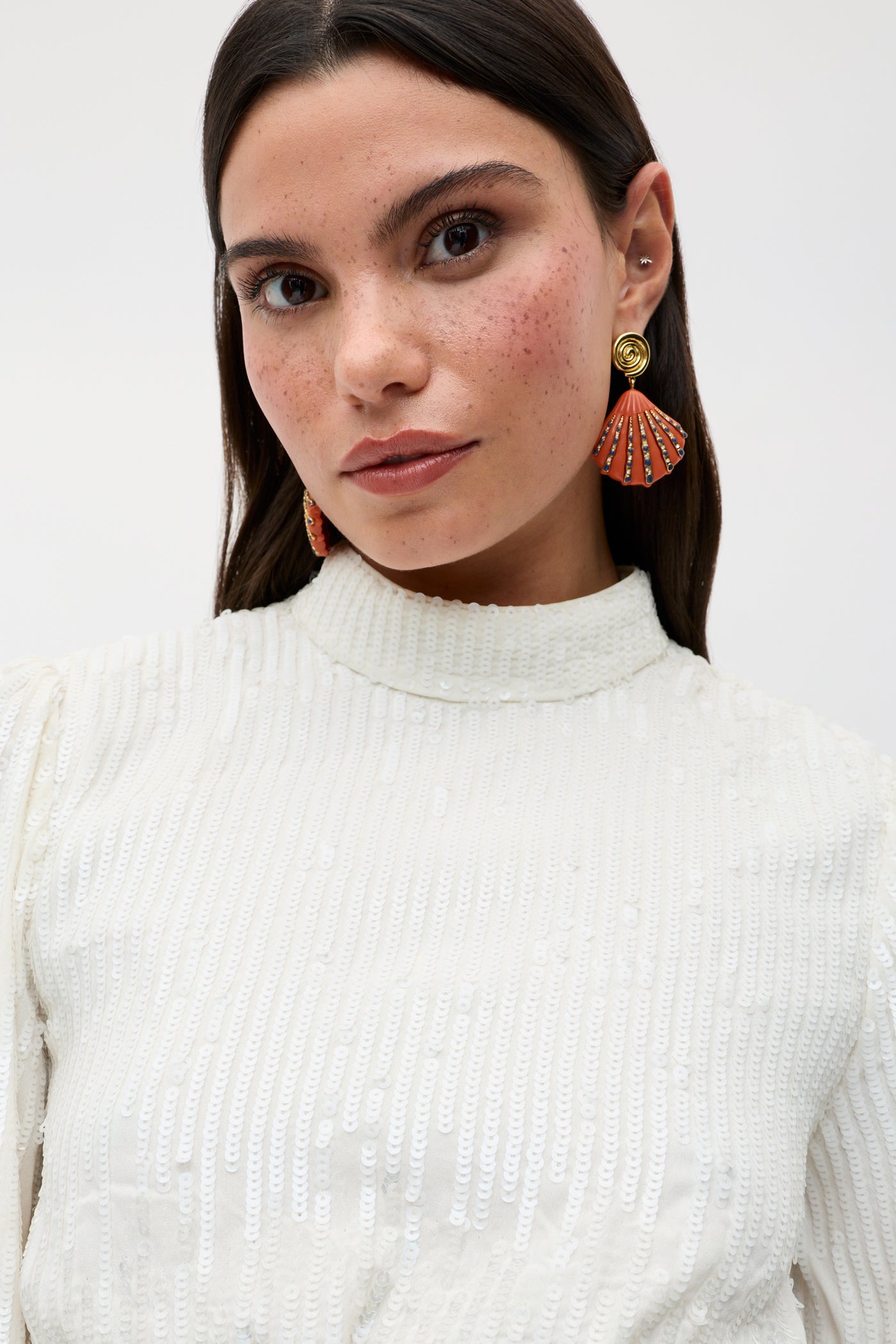 A woman with long dark hair, wearing the lara - ivory black bow dress—a white textured piece featuring a tie-back detail—and orange fan-shaped earrings, gazes directly at the camera with natural makeup and visible freckles against a plain light background.