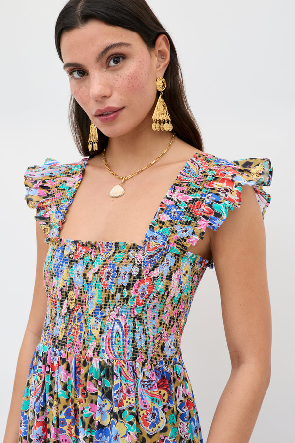 A woman with long brown hair wears the Miray - Gold dangling earrings along with a colorful, floral-patterned dress featuring ruffled sleeves and a shell necklace, standing against a plain light background.