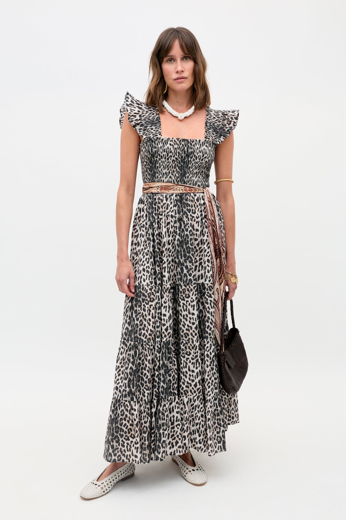 A woman poses against a white background wearing the Kendall - Nina Leopard Mocha dress, styled with a beaded necklace, gold bangles, a brown woven bag, and white slip-on shoes.