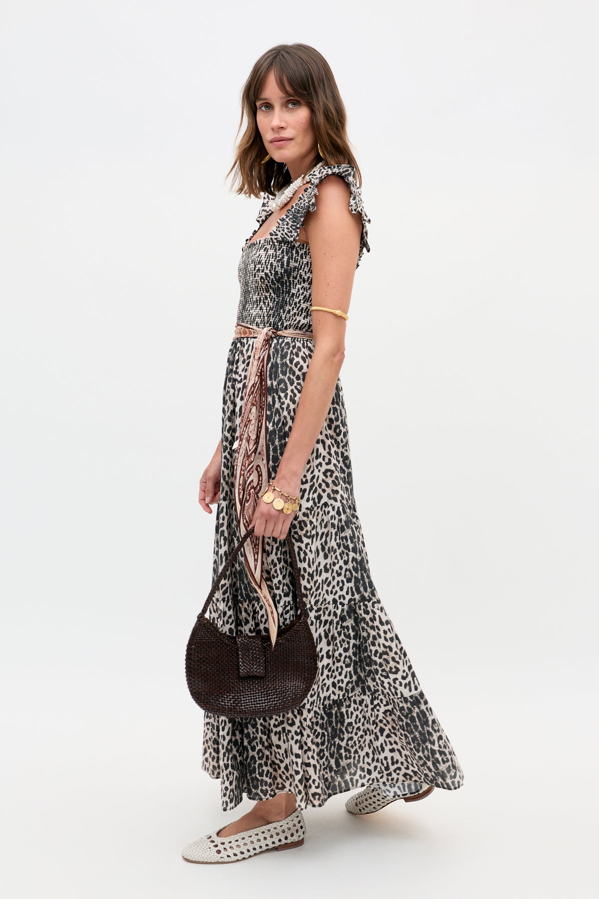 A woman poses sideways against a white background in the Kendall - Nina Leopard Mocha sleeveless ruffled maxi dress, carrying a round woven dark bag, wearing bracelets and patterned flats. Her wavy hair cascades over her shoulder.