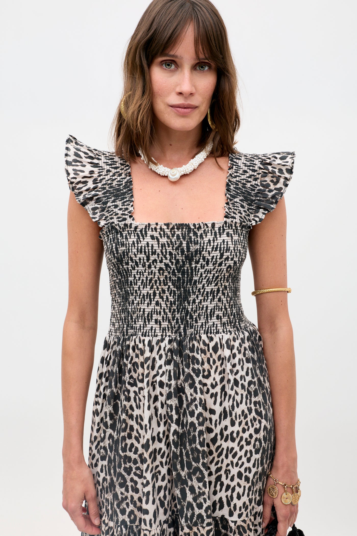 A woman with shoulder-length brown hair wears the Kendall - Nina Leopard Mocha dress with a ruffled, square neckline, accessorized with gold jewelry and a white shell necklace, standing against a plain backdrop.