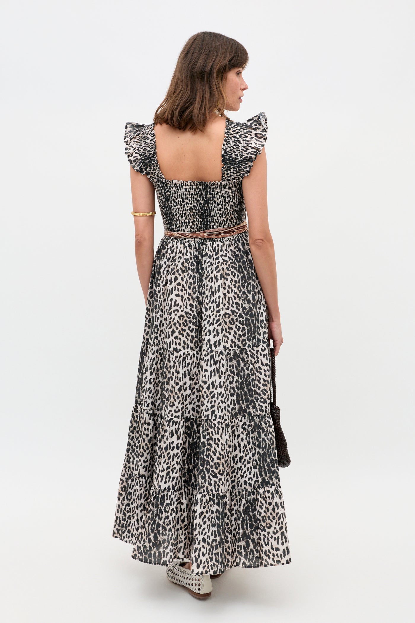 A woman faces away, wearing the Kendall - Nina Leopard Mocha ruffled sleeveless ankle-length dress. She carries a small brown bag and wears white cutout shoes against a plain white background.