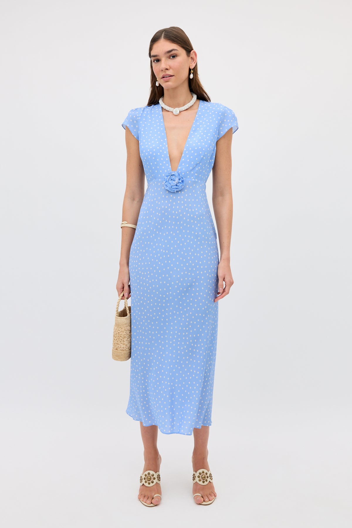 A woman wearing the Amy - Layla Dot Light Blue dress, featuring light blue polka dots, short sleeves, a deep V-neckline, and floral details, holds a straw bag while standing against a plain white background.