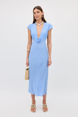 A woman wearing the Amy - Layla Dot Light Blue dress, featuring light blue polka dots, short sleeves, a deep V-neckline, and floral details, holds a straw bag while standing against a plain white background.