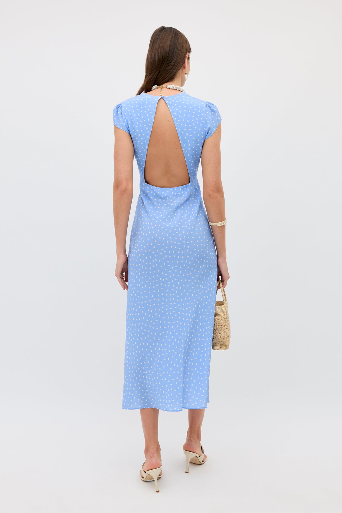 A woman wears the Amy - Layla Dot Light Blue dress, adorned with playful polka dots, short sleeves, and an open-back cutout. She stands facing away while holding a woven purse, styled with heeled sandals and gold accessories against a plain light background.