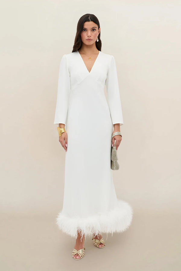 Wearing the Mya - Ivory Feathers dress—a long, bias-cut silk gown in white with a V-neckline and elegant ivory-feather trim at the hem—a woman stands against a light plain background. She accessorizes with gold jewelry, heeled sandals, and holds a textured clutch.
