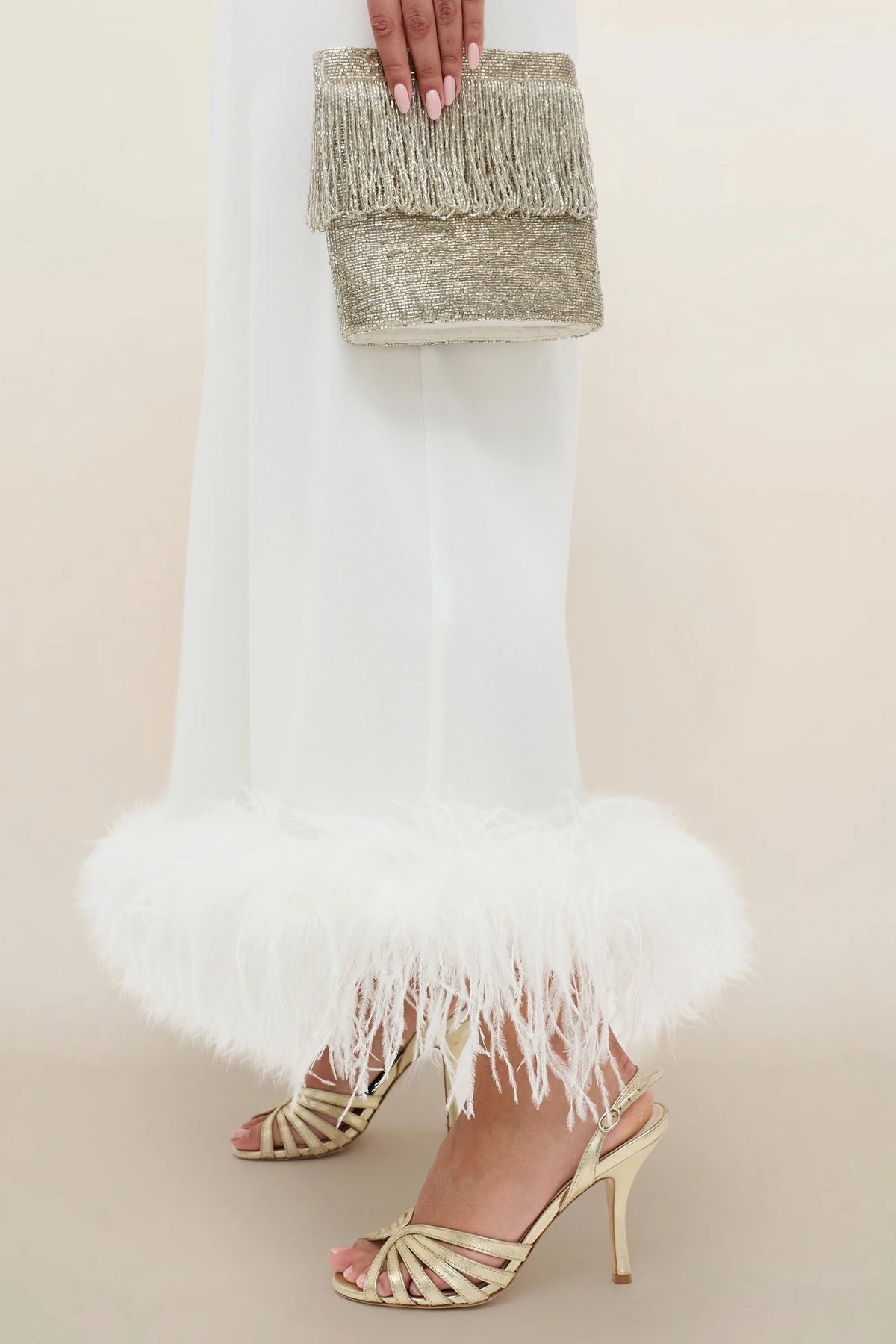 A person in the Mya - Ivory Feathers skirt holds a gold beaded clutch and wears gold high-heeled sandals with criss-cross straps.