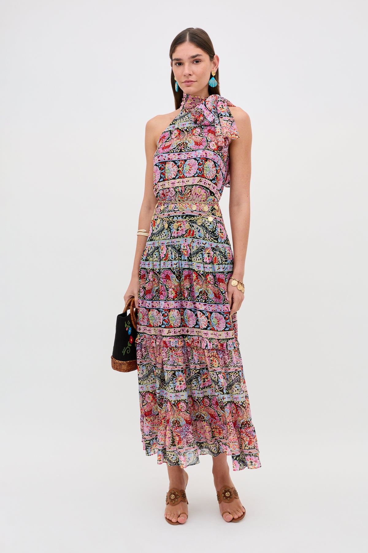 A woman poses against a white background wearing the Abi - Cara Stripe Black maxi dress, featuring colorful stripes, a sleeveless design, high neck, and ruffled tiers. She accessorizes with turquoise earrings, bracelets, sandals, and a black handbag.