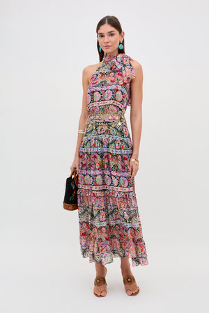 A woman poses against a white background wearing the Abi - Cara Stripe Black maxi dress, featuring colorful stripes, a sleeveless design, high neck, and ruffled tiers. She accessorizes with turquoise earrings, bracelets, sandals, and a black handbag.