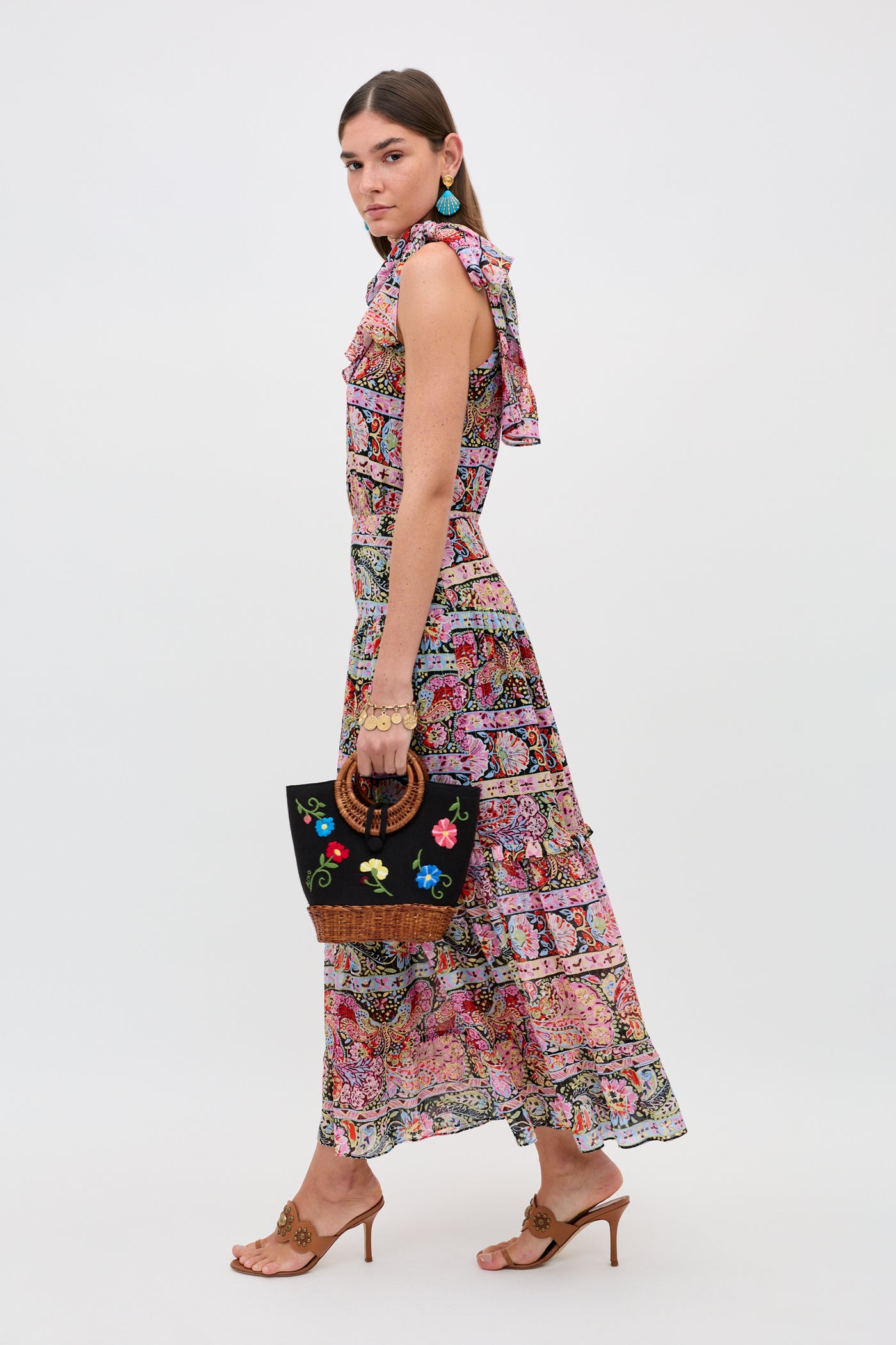 A woman wears the Abi - Cara Stripe Black maxi dress, styled with tan heeled sandals, turquoise earrings, and a black floral embroidered handbag, standing against a plain white background.