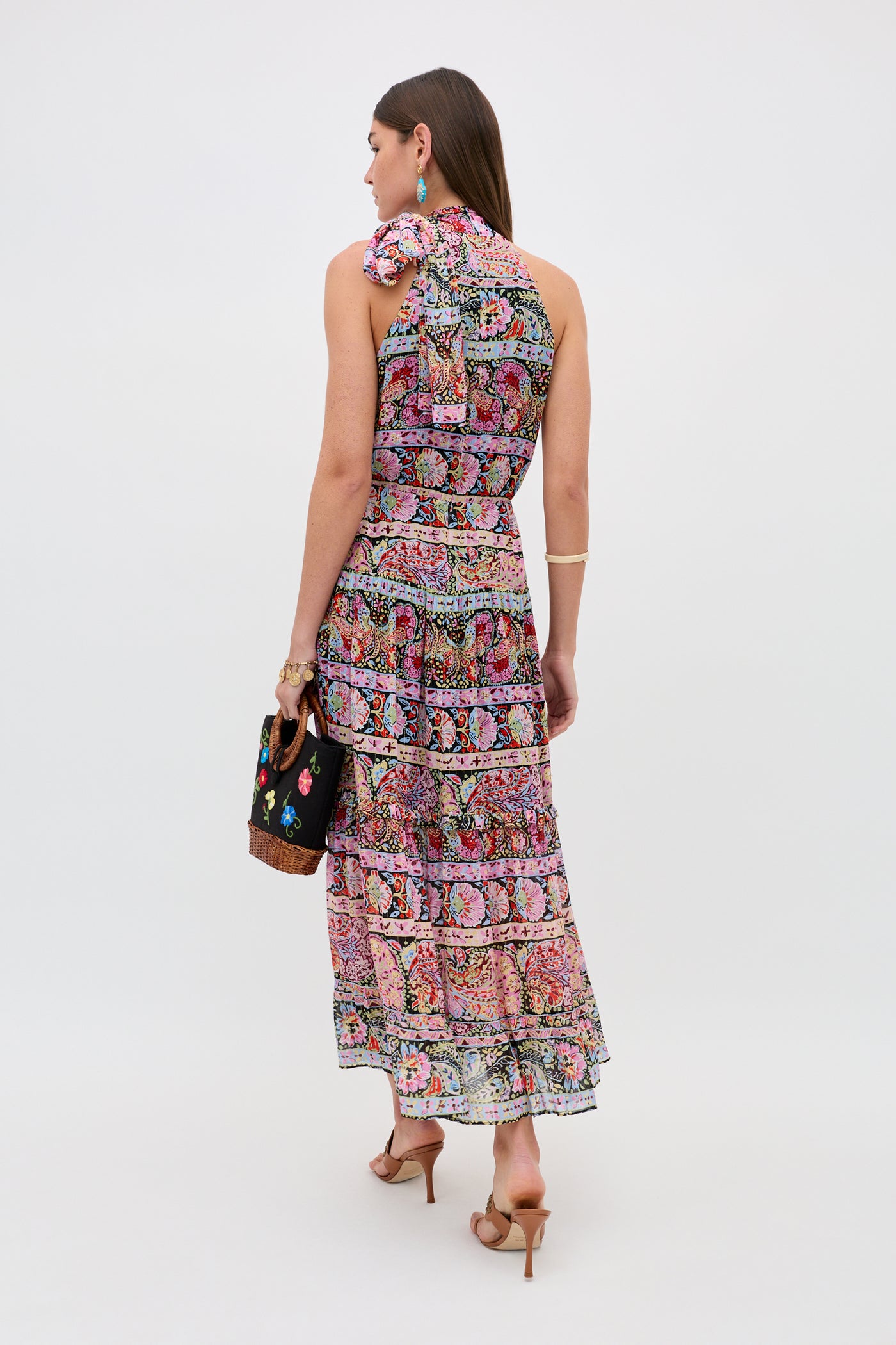A woman faces away, wearing the Abi - Cara Stripe Black sleeveless maxi dress with a tiered skirt and bow shoulder detail. She pairs it with a black floral handbag, heels, earrings, and bracelets against a plain white background.