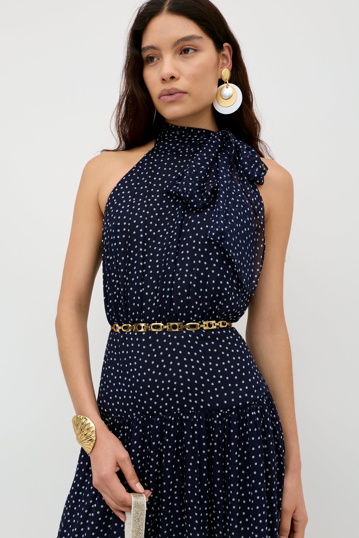 A woman wears the Abi - Mini Navy Polka Dot dress, a sleeveless georgette design with white spots, high bow neckline, gold chain belt, statement earrings, and cuff—perfect for special occasions—posed against a plain light backdrop.