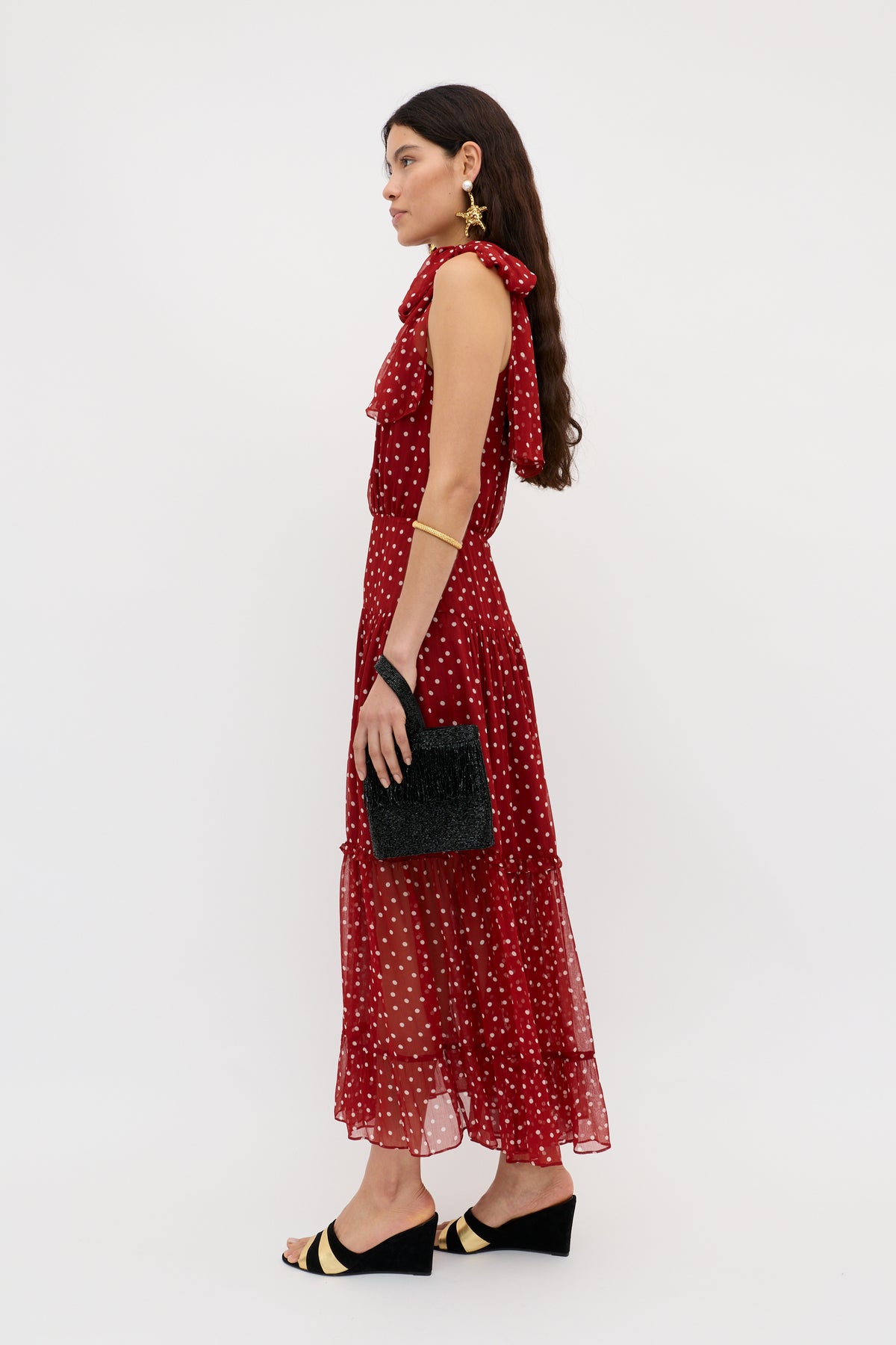 A woman in the Abi - Layla Dot Red sleeveless midi dress and black wedge sandals stands in profile, holding a textured black clutch. She has long brown hair, gold jewelry, and is posed against a plain white background.