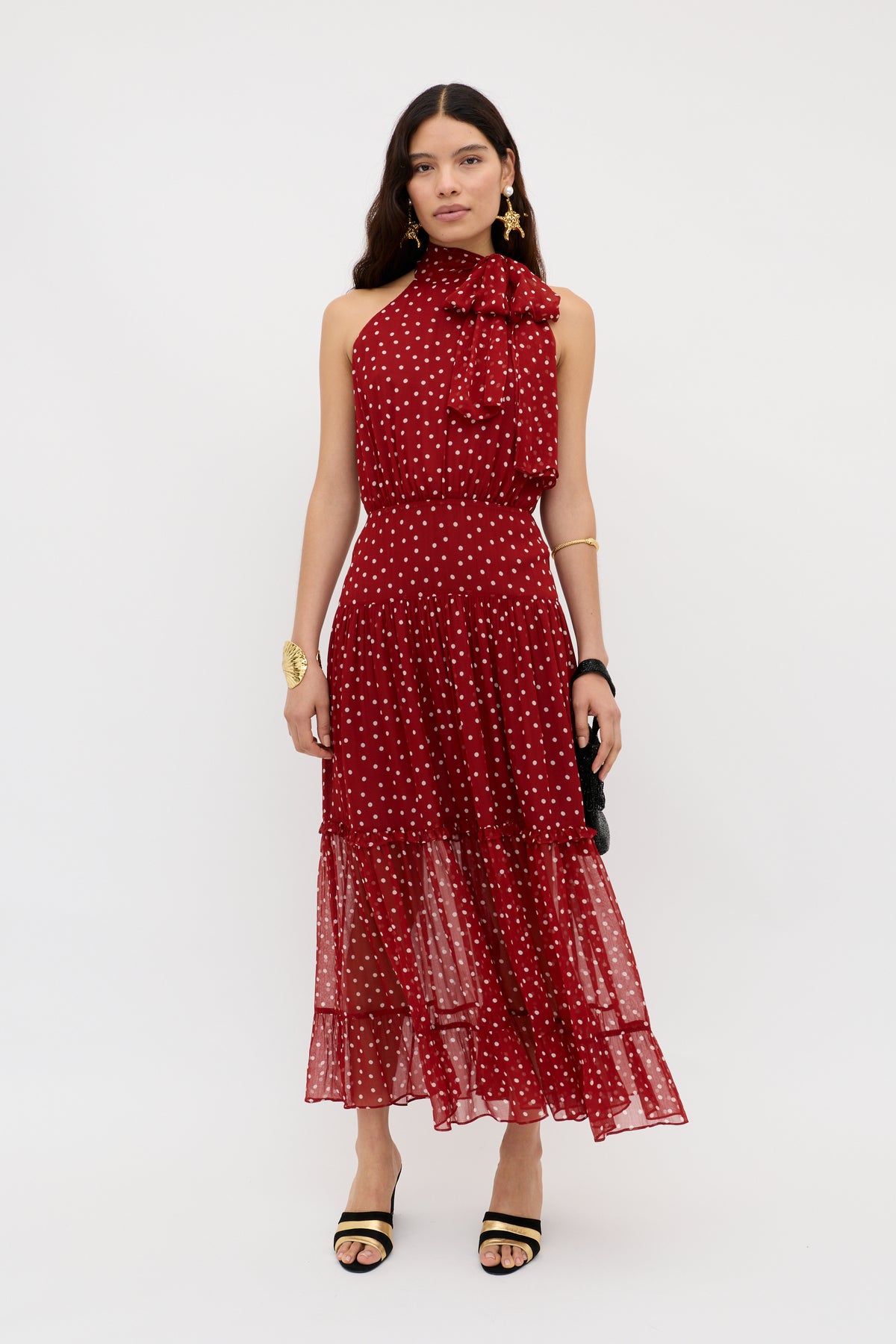 A woman stands against a white background wearing the Abi - Layla Dot Red midi dress in crinkled chiffon with a sleeveless halter-neck, ruffled neckline, and tiered skirt, styled with statement jewelry and black heeled sandals.