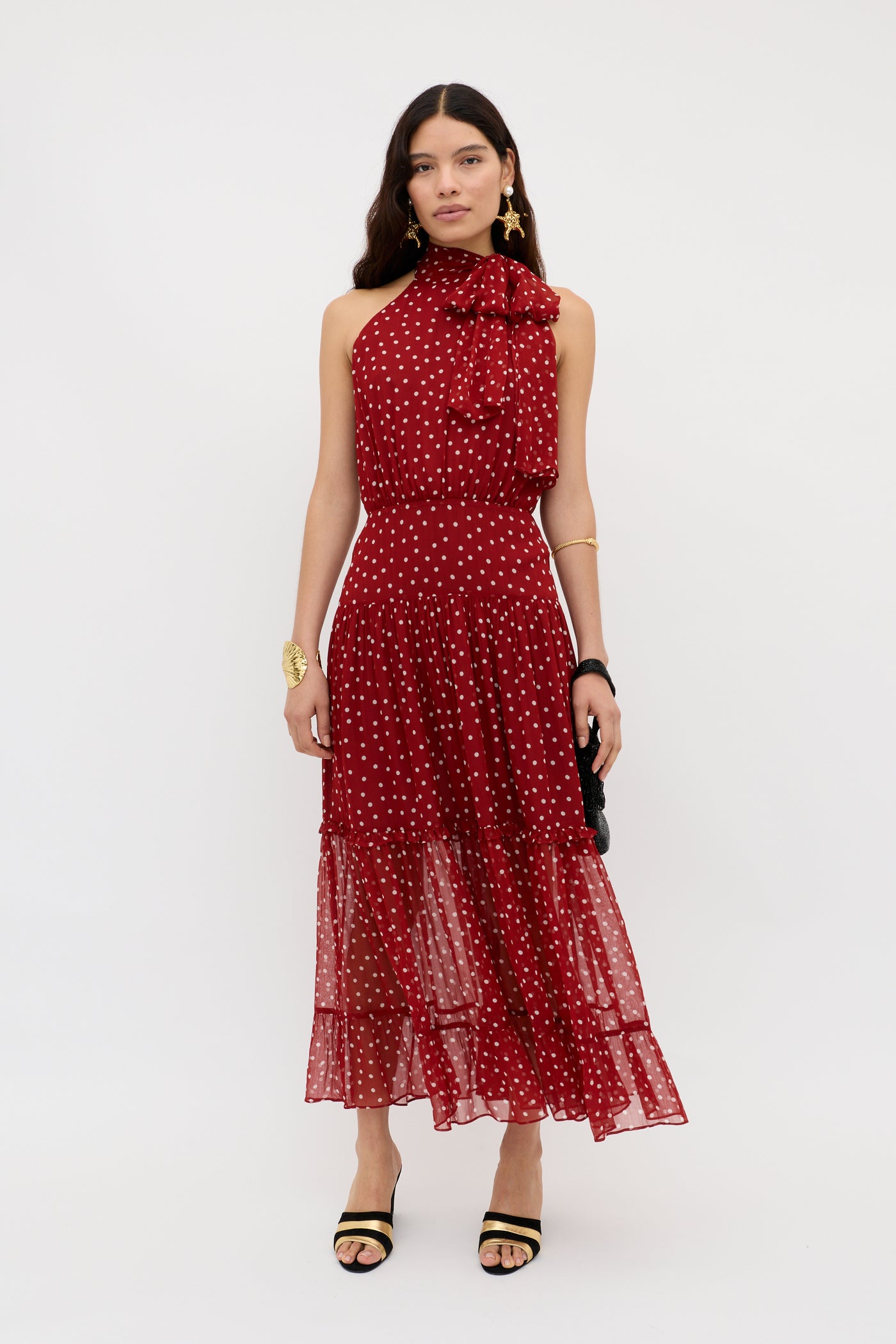 A woman stands against a white background wearing the Abi - Layla Dot Red midi dress in crinkled chiffon with a sleeveless halter-neck, ruffled neckline, and tiered skirt, styled with statement jewelry and black heeled sandals.