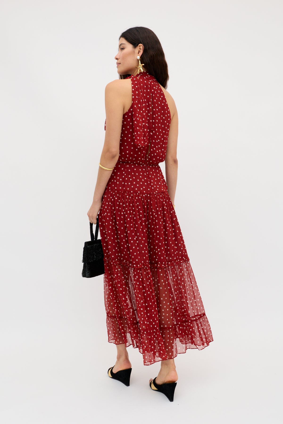 A woman wears the Abi - Layla Dot Red midi dress, sleeveless and ankle-length in crinkled red chiffon with white polka dots. Facing away, she holds a black handbag, wears black heels, has dark hair down and gold accessories.