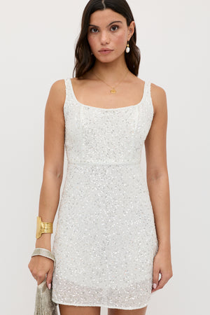 A woman wearing the ronan - squiggle sequin ivory sleeveless mini dress with a square neckline, accessorized with gold jewelry and a silver fringed clutch, stands against a plain light background.