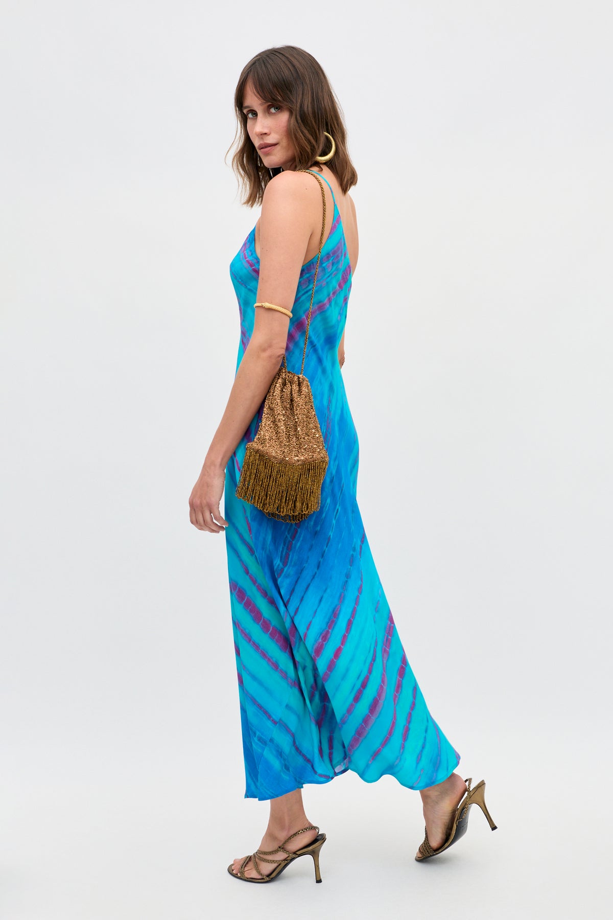 A woman wearing the Doutzen - Tie Dye Blue maxi dress, paired with gold hoop earrings, strappy heels, and a brown textured shoulder bag, stands against a plain light background.