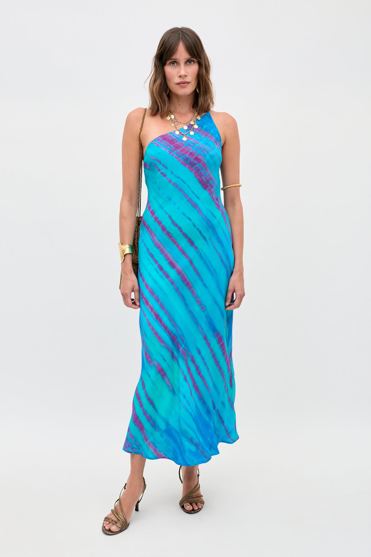 A woman wears the Doutzen - Tie Dye Blue one-shoulder dress, styled with gold jewelry and heeled sandals, standing against a plain light background.