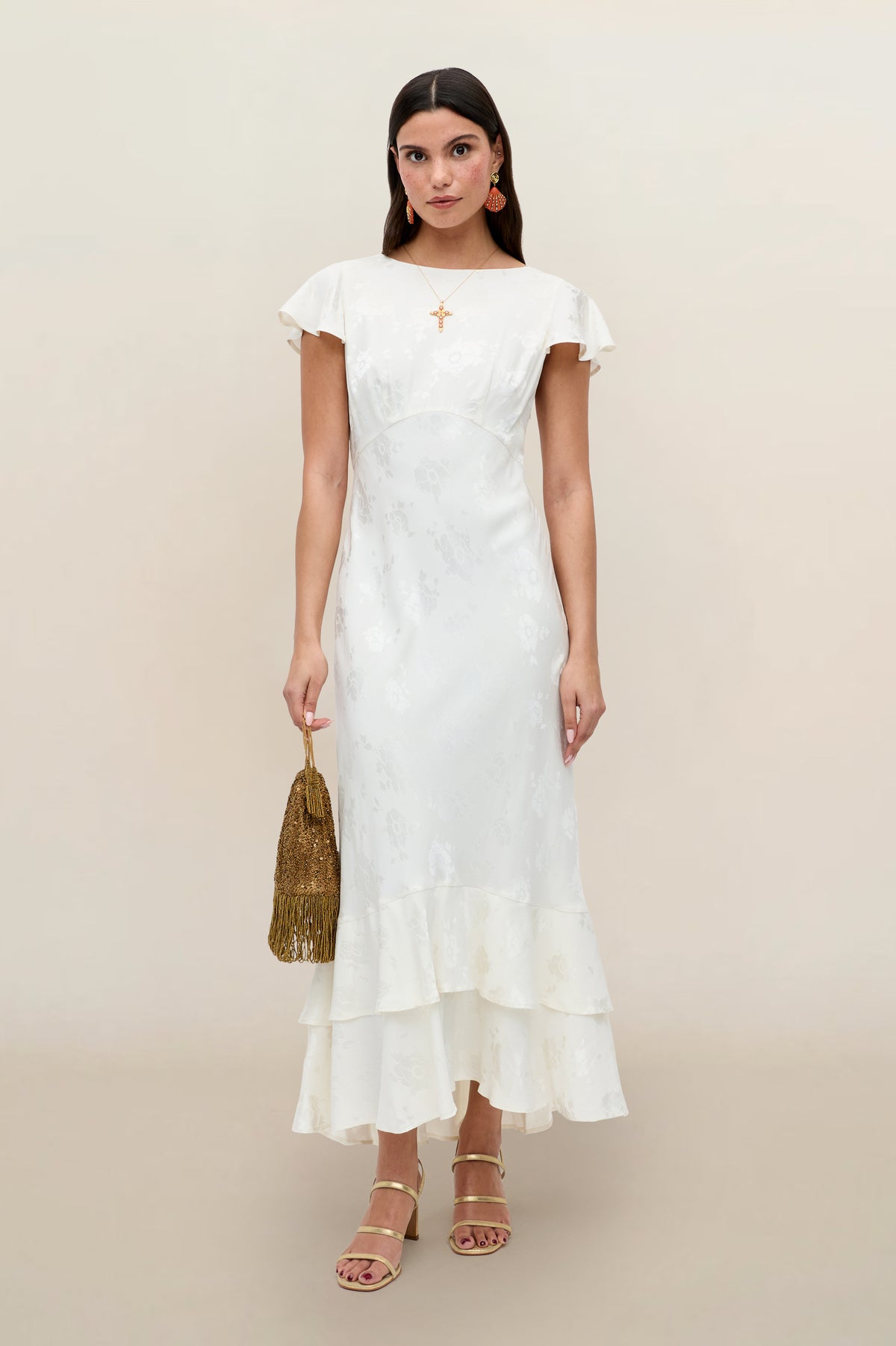 A woman wears the Liberty - Poppy ivory jacquard ankle-length dress with ruffle hem and cap sleeves, styled with gold earrings, strappy heels, and a gold fringed bag against a plain beige background.