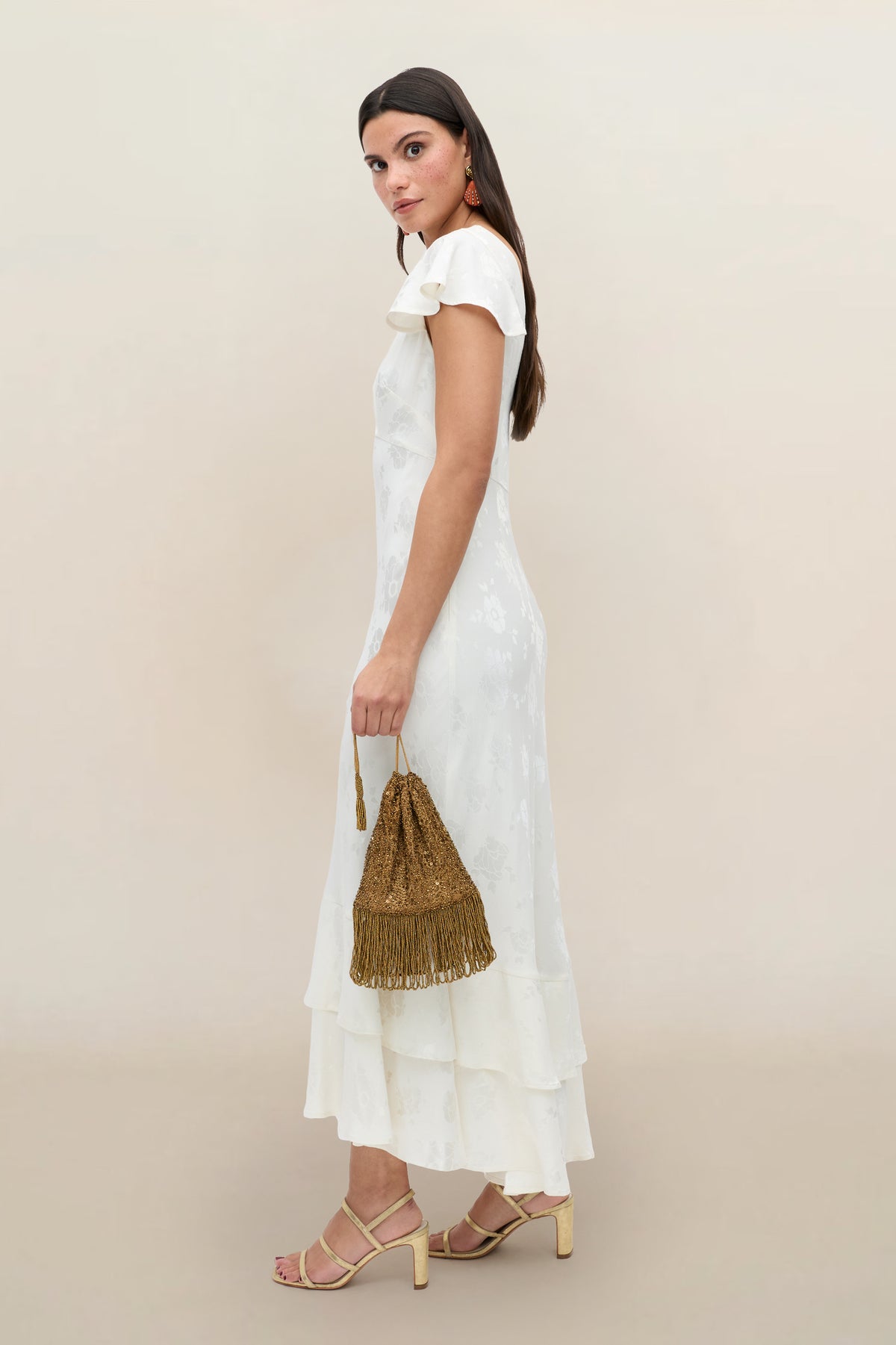 A woman in the Liberty - Poppy ivory jacquard, ruffled, ankle-length vintage wedding dress with cap sleeves stands sideways holding a gold fringed handbag, wearing heeled sandals and looking neutrally at the camera against a beige background.