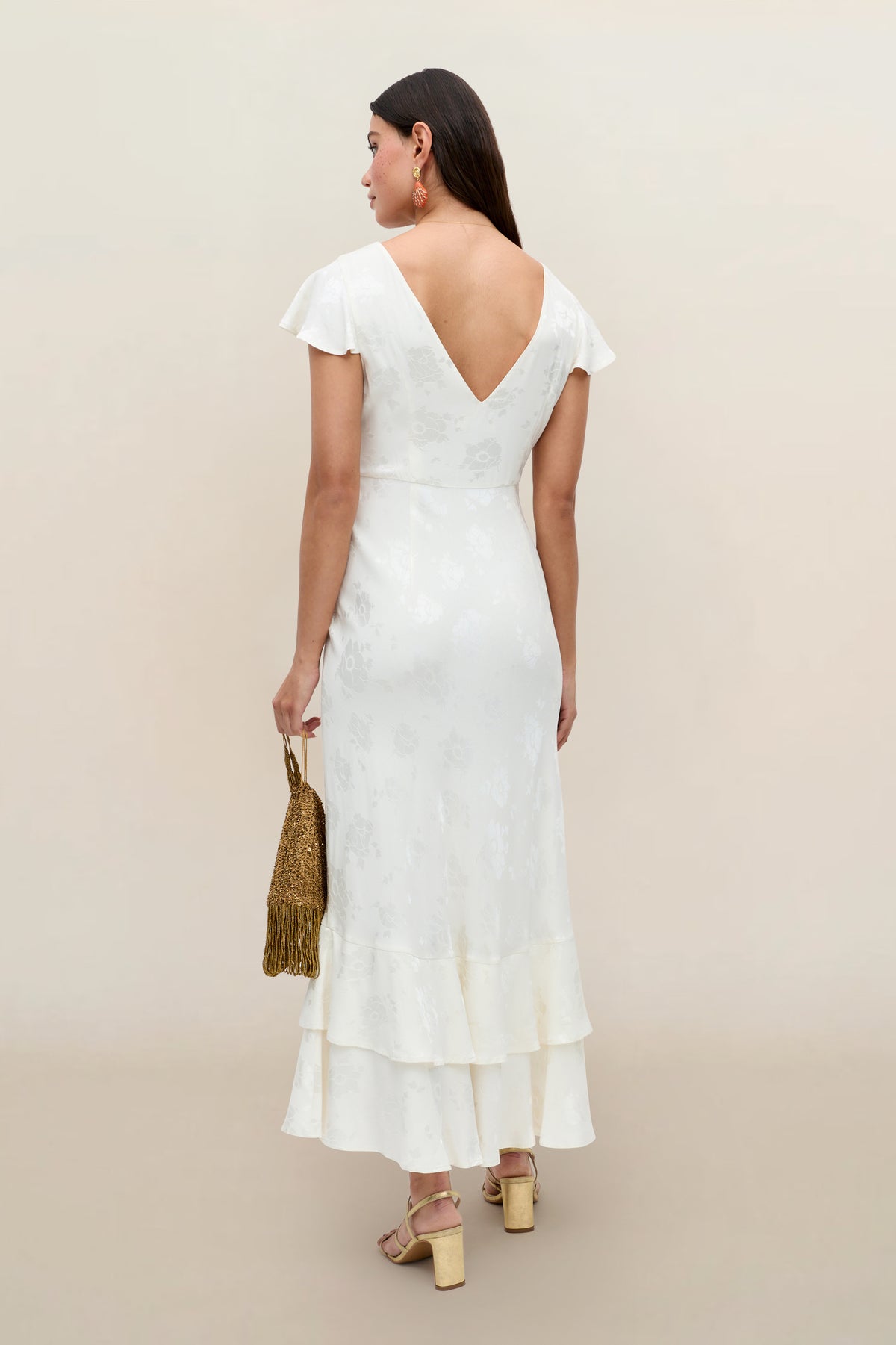 A woman faces away in the Liberty - Poppy ivory jacquard midi dress with cap sleeves, V-back, and ruffled hem. She carries a gold beaded handbag and wears chunky heeled sandals against a light beige background.