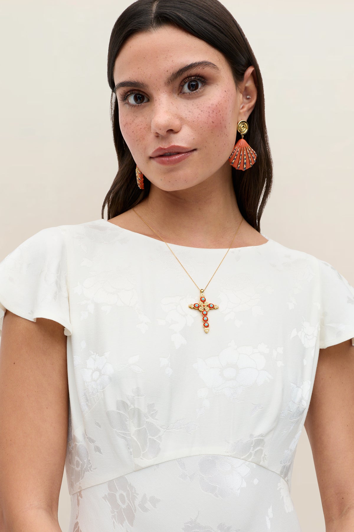 A woman wears the Liberty - Poppy ivory jacquard floral-patterned dress with cap sleeves, paired with orange fan earrings and a gold cross necklace. She has long brown hair and a neutral expression against a plain background.