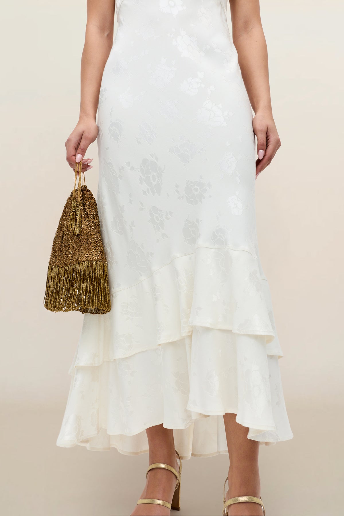 A woman wears the Liberty - Poppy, an ivory jacquard floral ruffled midi dress, styled with a gold beaded fringe handbag and gold strappy heels. The image is cropped from shoulders to feet.