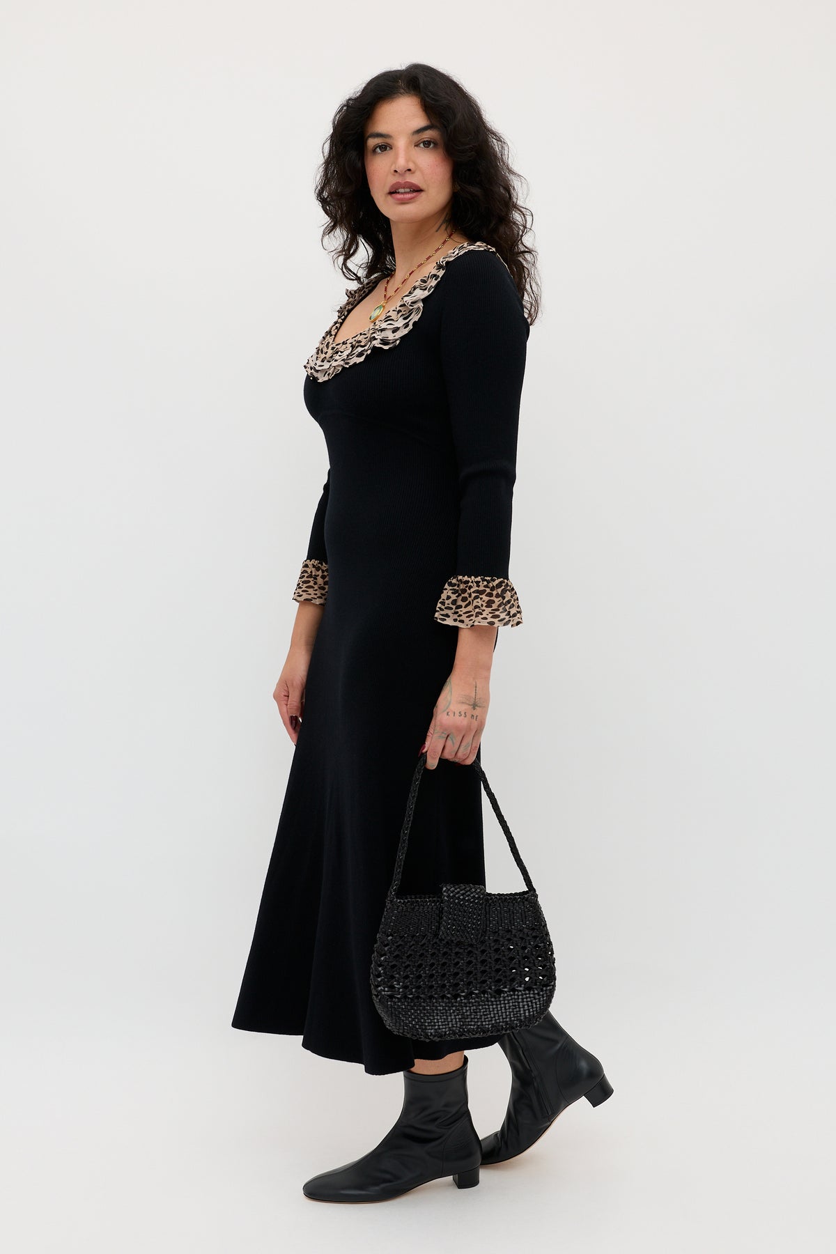 A woman with curly dark hair wears the Mika - Antelope Spot Black merino wool dress with ruffled leopard trim on the neckline and cuffs, black ankle boots, and holds a black woven handbag against a white background.