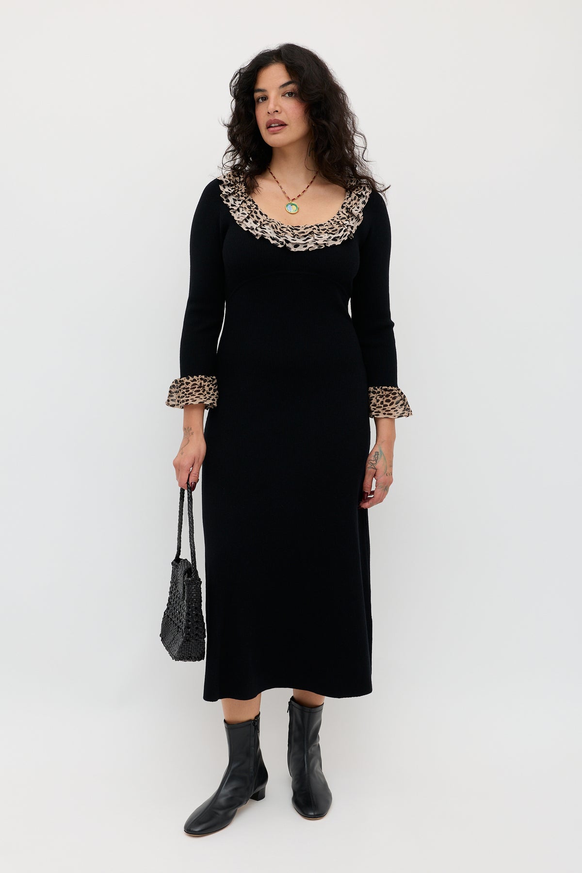 A woman stands against a white background wearing the Mika - Antelope Spot Black merino wool dress with ruffled leopard trim, black ankle boots, and holding a textured black handbag.