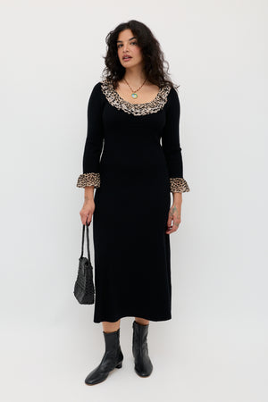 A woman stands against a white background wearing the Mika - Antelope Spot Black merino wool dress with ruffled leopard trim, black ankle boots, and holding a textured black handbag.