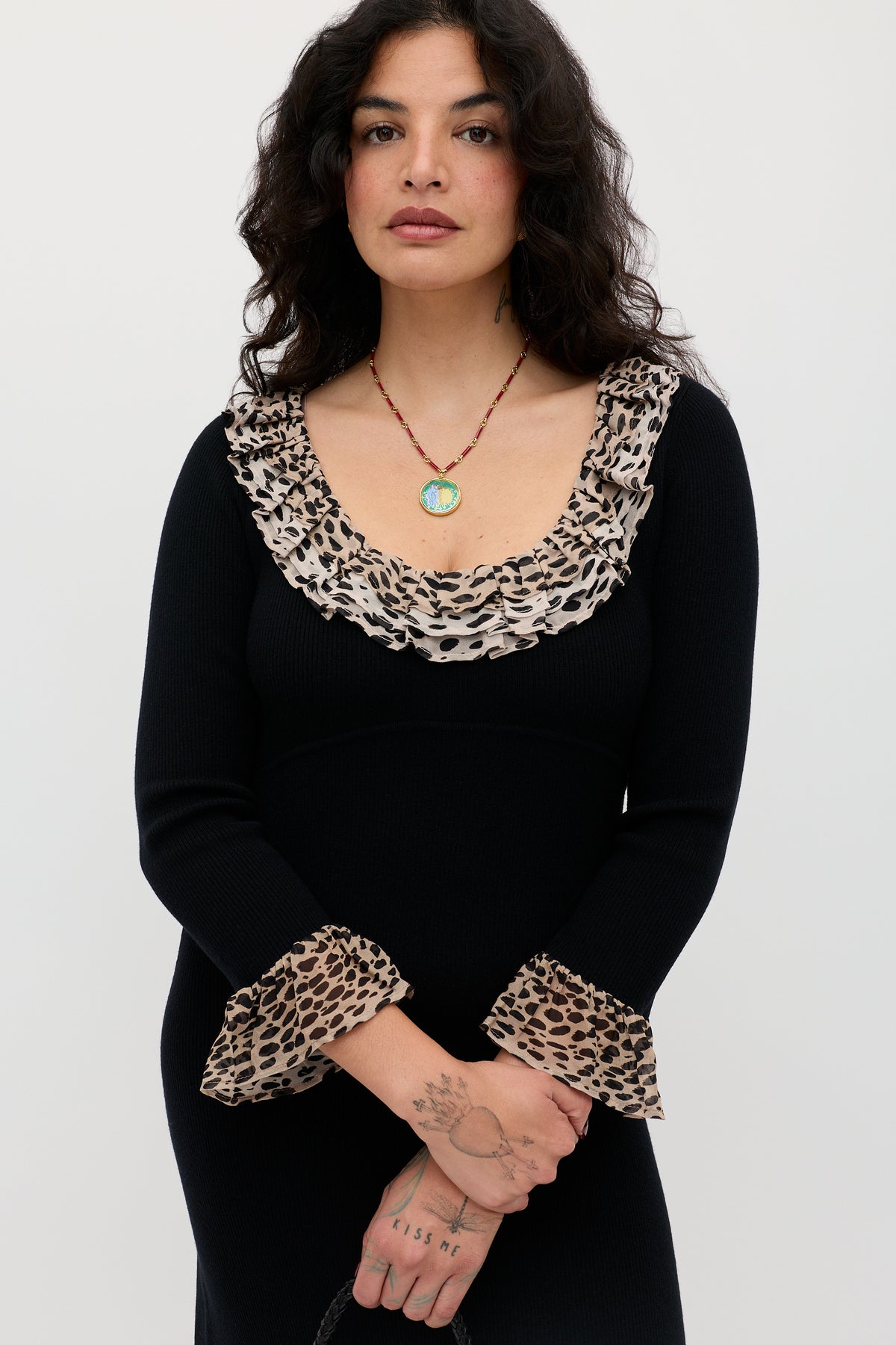 A woman with wavy dark hair wears the Mika - Antelope Spot Black dress, featuring a scoop neck and ruffled leopard trim on the neckline and cuffs. She stands against a plain backdrop, accessorized with a beaded necklace and visible hand tattoos.