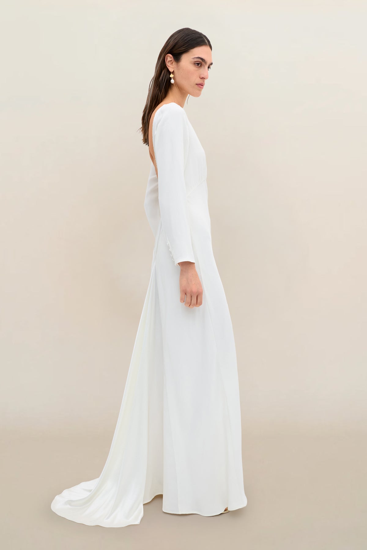 A woman stands in profile wearing the Cesca - Ivory, a vintage-inspired, long white elegant dress with long sleeves and a satin train, against a plain light beige background.
